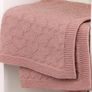 May include: A dusty rose knitted blanket featuring a textured geometric diamond pattern across the center and a wide, ribbed border along the edges. The fabric displays a consistent knit stitch throughout, creating a soft and uniform appearance.
