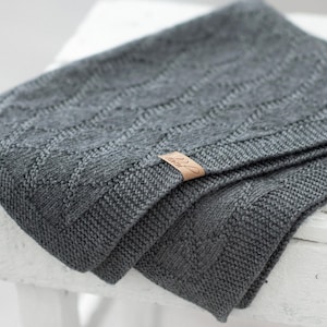 Puede incluir: Una manta gris tejida, doblada, con un patrón de diamantes y un borde liso. Una pequeña etiqueta rectangular de cuero está adherida a la manta. La manta está sobre un taburete de madera blanco desgastado.