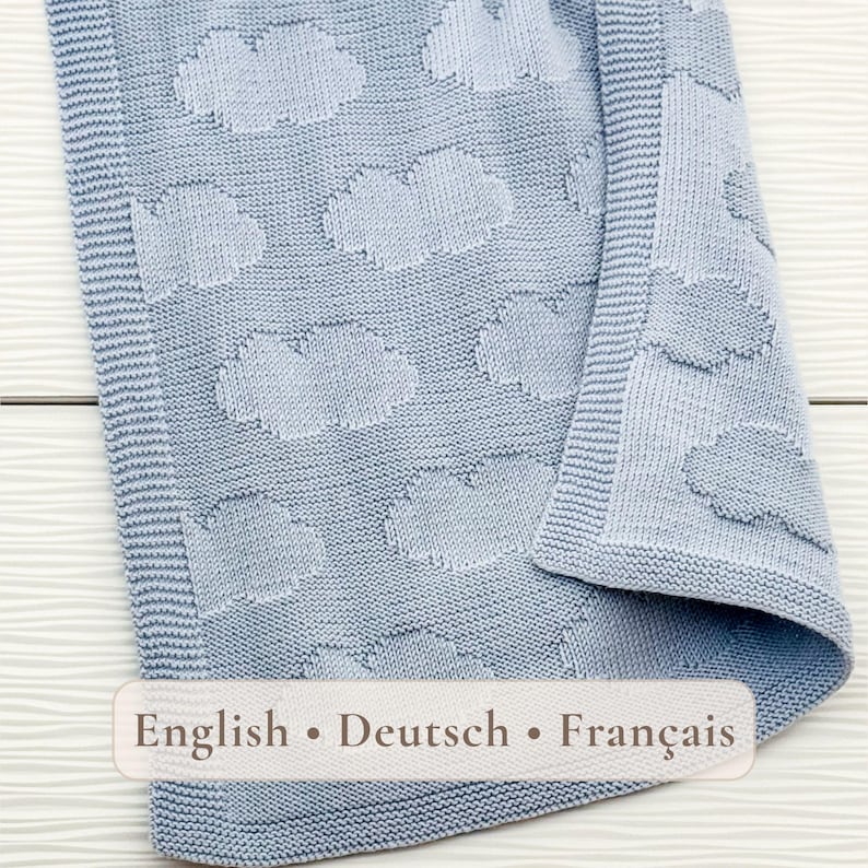 May include: A light blue knitted blanket featuring a repeating pattern of clouds. The fabric has a textured knit surface with a distinct border along the edge. The text "English &bull; Deutsch &bull; Fran&ccedil;ais" is printed in a horizontal banner across the bottom.