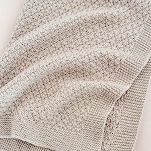 Puede incluir: Una manta de punto plegada, de tonos neutros, con un patrón texturizado. La manta presenta un diseño de puntada detallado y un borde acabado. El color es una mezcla de tonos neutros claros y oscuros, creando un contraste sutil. La manta parece suave y acogedora.