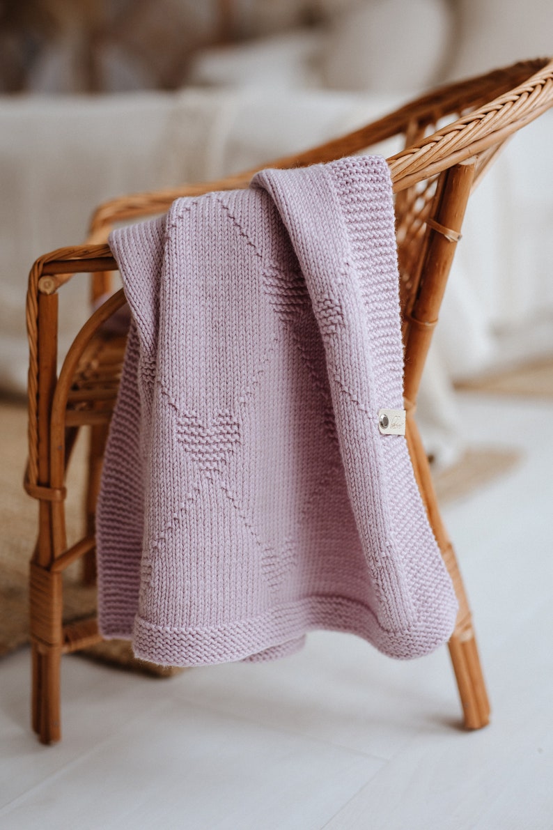 K&ouml;nnte beinhalten: Eine fliederfarbene Strickdecke mit Herzmuster, die &uuml;ber einem Korbstuhl drapiert ist. Die Decke hat ein strukturiertes Aussehen und ein kleines angebrachtes Etikett. Der Stuhl ist aus hellbraunem Korbgeflecht.