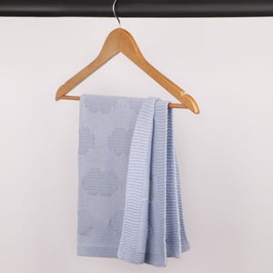 May include: A light blue knitted blanket featuring a repeating pattern of textured clouds. The blanket is draped over a wooden clothes hanger, showcasing a ribbed border along the edges and a soft, consistent knit construction throughout the fabric.