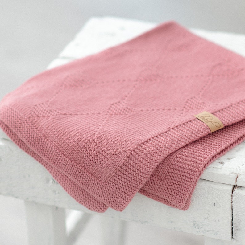 K&ouml;nnte beinhalten: Eine gefaltete, altrosa gestrickte Decke mit einem Rautenmuster. Die Decke hat einen gerippten Rand und ein kleines, rechteckiges Etikett mit kursivem Text. Die Decke ist auf einer wei&szlig;en Oberfl&auml;che gefaltet.