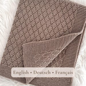 May include: A light brown knitted blanket featuring a textured diamond pattern across the center and a solid garter stitch border along the edges. The fabric displays a consistent knit construction with a folded corner revealing the reverse side.