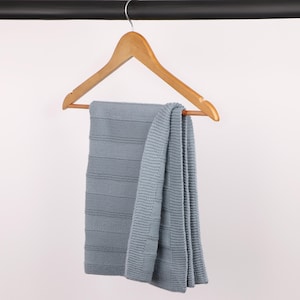 May include: A light blue knitted blanket with a textured horizontal stripe pattern and a solid border, draped over a wooden clothes hanger. The blanket features a consistent knit construction throughout, showcasing a soft, muted blue color tone.
