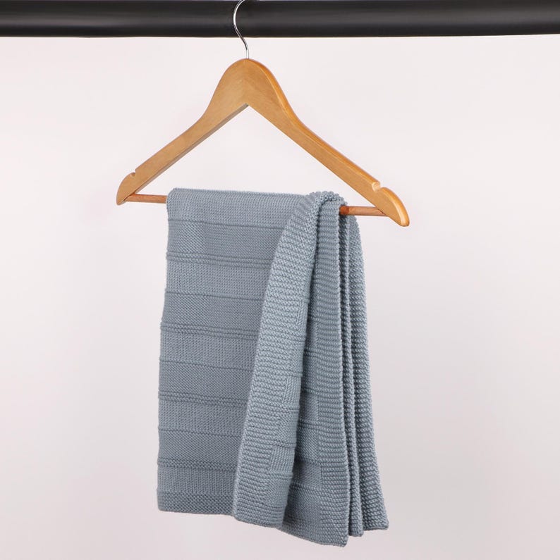 May include: A light blue knitted blanket with a textured horizontal stripe pattern and a solid border, draped over a wooden clothes hanger. The blanket features a consistent knit construction throughout, showcasing a soft, muted blue color tone.