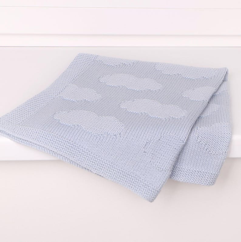 May include: A light blue knitted baby blanket featuring a repeating pattern of soft, textured clouds. The blanket has a distinct border with a fine knit stitch, while the central area displays the cloud motifs in a slightly raised, knit texture.