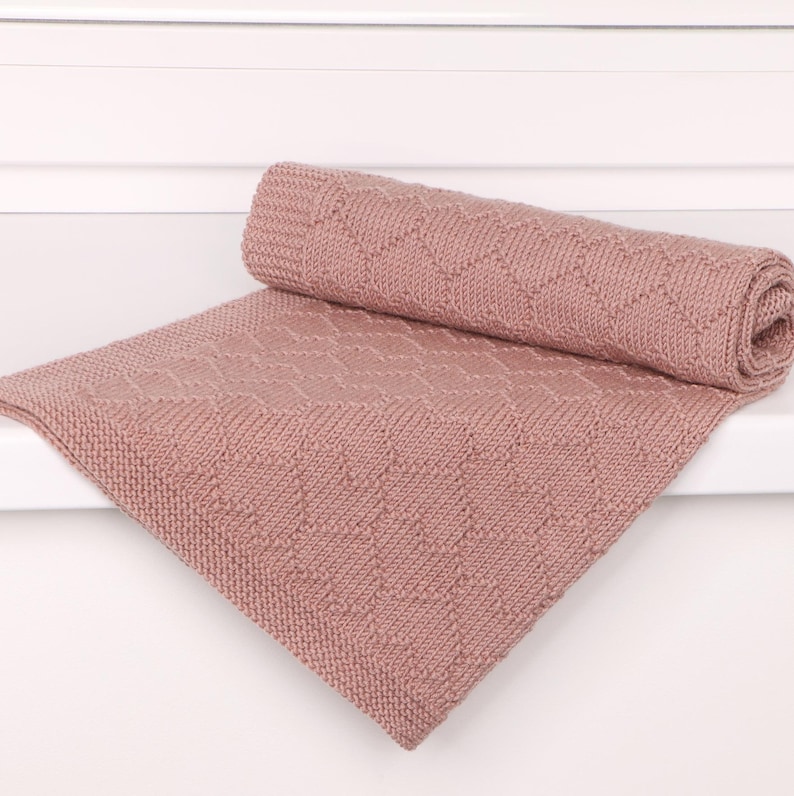 May include: A dusty rose knitted blanket featuring a repeating diamond pattern throughout the fabric. The blanket is folded over itself, showcasing a textured knit construction with a consistent border along the edges of the soft pink textile.