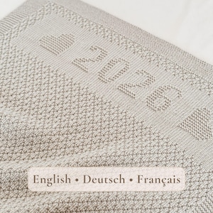 May include: A light beige knitted blanket featuring a textured diamond pattern. The border displays the text 2026 flanked by two heart shapes, all integrated into the knit. The fabric has a consistent weave and a soft, neutral color tone.
