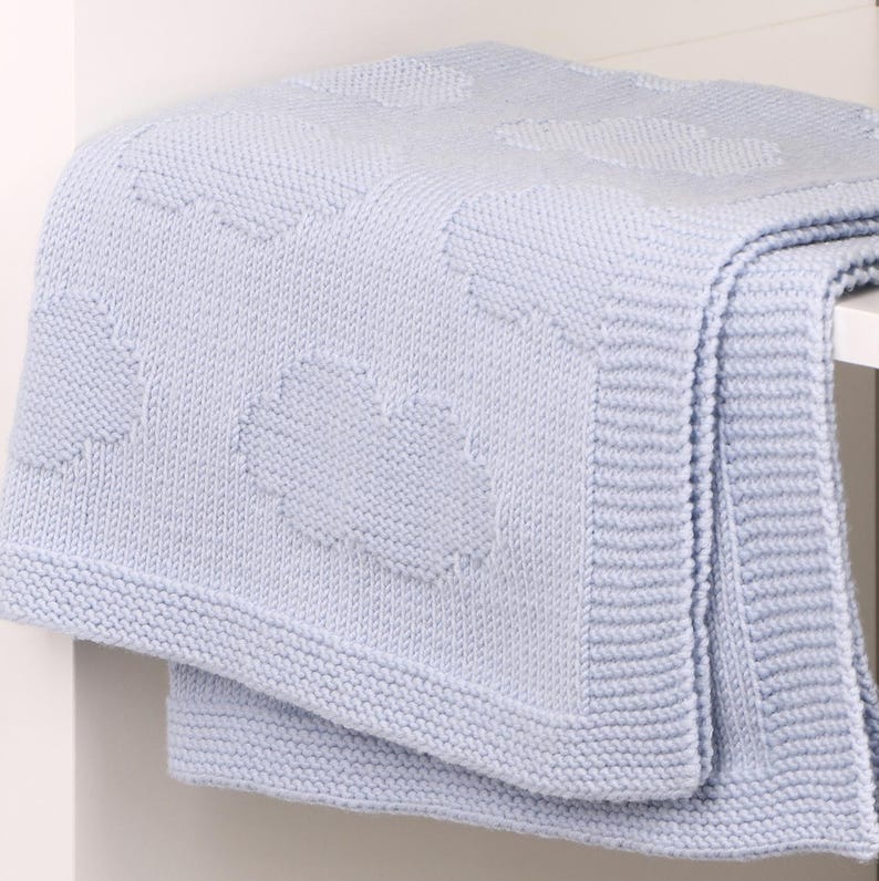 May include: A light blue knitted blanket featuring a repeating pattern of textured clouds. The blanket has a wide, ribbed border along the edges and a soft, consistent knit construction throughout the entire piece, showcasing a uniform color.
