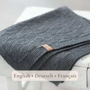 Puede incluir: Una manta de punto gris oscuro con un patrón de cable de diamante texturizado y un borde acanalado. Una pequeña etiqueta rectangular de color cuero tostado con texto en cursiva está unida al borde de la tela, añadiendo un detalle sutil al diseño.