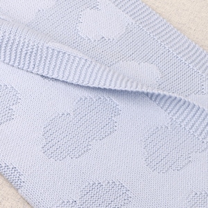 May include: A light blue knitted blanket featuring a repeating pattern of raised, textured cloud shapes. The fabric has a consistent garter stitch texture throughout, with a folded edge revealing the underside of the soft, woven material.