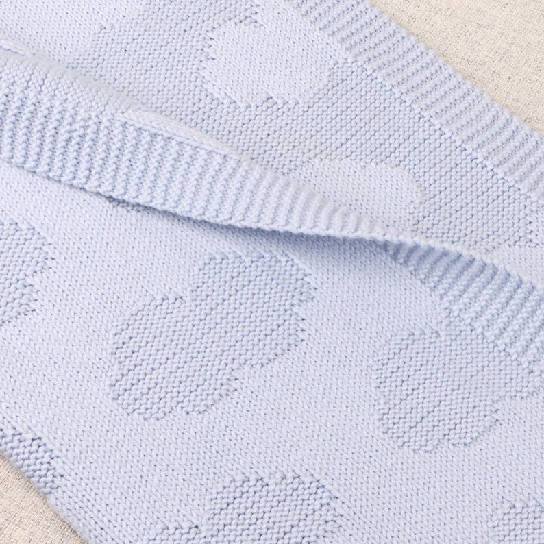 May include: A light blue knitted blanket featuring a repeating pattern of raised, textured cloud shapes. The fabric has a consistent garter stitch texture throughout, with a folded edge revealing the underside of the soft, woven material.