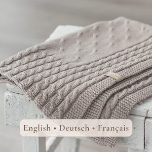 May include: A light beige knitted blanket featuring a textured pattern of vertical cable knit stripes and small raised dots. The fabric has a soft appearance with a small rectangular label attached to the edge. The text English • Deutsch • Français is visible.