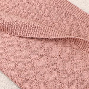 May include: A dusty rose knitted blanket featuring a repeating geometric honeycomb pattern. The fabric displays a textured knit construction with a distinct ribbed border edge, showcasing a consistent soft pink color throughout the entire textile piece.