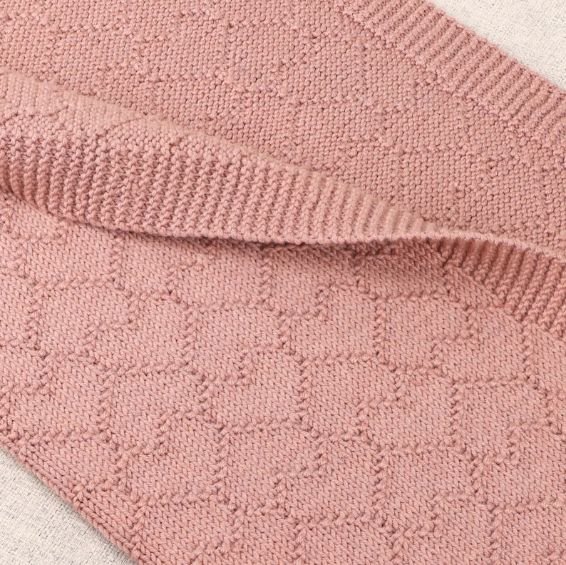 May include: A dusty rose knitted blanket featuring a repeating geometric honeycomb pattern. The fabric displays a textured knit construction with a distinct ribbed border edge, showcasing a consistent soft pink color throughout the entire textile piece.