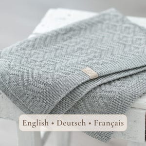 Könnte beinhalten: Eine hellgraue Strickdecke mit einem texturierten geometrischen Rautenmuster auf dem gesamten Stoff. Ein kleines, rechteckiges beiges Stoffetikett mit kursivem Text ist am Rand der Decke befestigt, die in sich gefaltet ist.