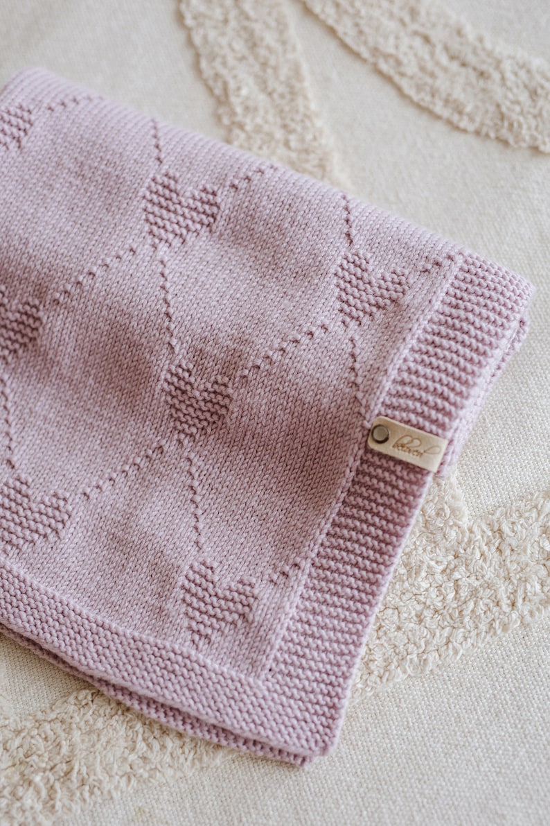 K&ouml;nnte beinhalten: Eine gestrickte Decke in Lavendel mit einem Herzmuster. Die Decke hat einen gerippten Rand und ein kleines Lederetikett. Die Decke ist aus weichem Material und f&uuml;r ein Baby oder Kind geeignet. Ma&szlig;e in Zentimetern.
