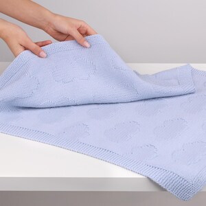 May include: A light periwinkle blue knitted blanket featuring a repeating textured cloud pattern across the surface. The blanket has a wide, garter stitch border along the edges, providing a consistent knit texture throughout the entire piece.