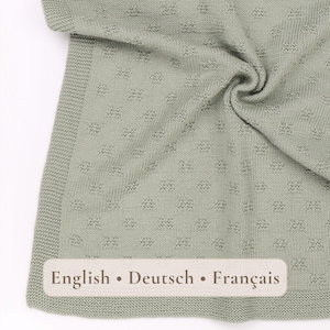 May include: A sage green knitted blanket featuring a repeating textured square pattern and a solid garter stitch border. A rectangular label is attached to the bottom corner with the text English • Deutsch • Français printed in dark brown letters.