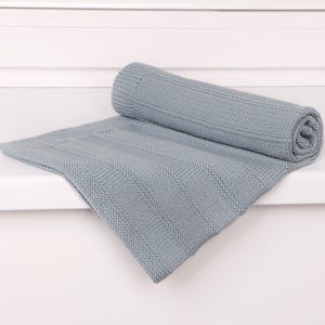 May include: A light blue knitted blanket featuring a textured pattern of alternating vertical garter stitch stripes and smooth stockinette stitch panels. The blanket is partially rolled up, showcasing the consistent weave and soft, matte finish of the fabric.