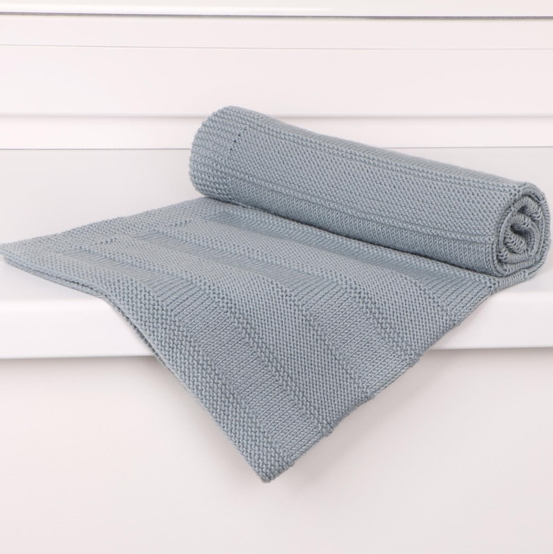 May include: A light blue knitted blanket featuring a textured pattern of alternating vertical garter stitch stripes and smooth stockinette stitch panels. The blanket is partially rolled up, showcasing the consistent weave and soft, matte finish of the fabric.