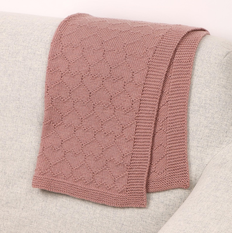 May include: A dusty rose knitted blanket featuring a repeating heart pattern throughout the main body and a wide, textured garter stitch border along the edges. The fabric has a consistent, soft-looking knit construction with a uniform color.