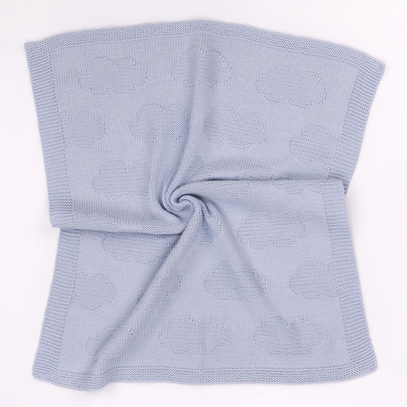May include: A light blue knitted blanket featuring a repeating pattern of textured clouds across the surface. The blanket has a wide, garter stitch border framing the central design, with a soft, uniform color throughout the entire piece.