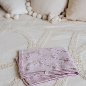 K&ouml;nnte beinhalten: Eine gefaltete, hellviolette Babydecke mit Rautenmuster und einem kleinen Etikett. Die Decke besteht aus einem weichen Material und liegt auf einer cremefarbenen Tagesdecke. Dekorative Kissen sind im Hintergrund.