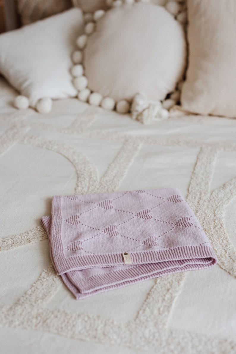 K&ouml;nnte beinhalten: Eine gefaltete, hellviolette Babydecke mit Rautenmuster und einem kleinen Etikett. Die Decke besteht aus einem weichen Material und liegt auf einer cremefarbenen Tagesdecke. Dekorative Kissen sind im Hintergrund.