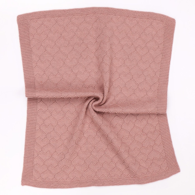 May include: A dusty rose knitted blanket featuring a repeating heart pattern throughout the center panel. The blanket is finished with a wide, textured garter stitch border along all four edges, creating a soft and uniform appearance.
