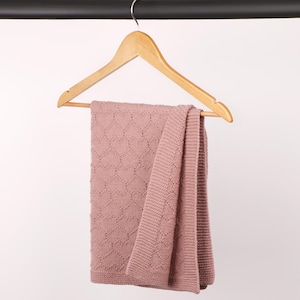 May include: A dusty rose knitted blanket featuring a repeating heart pattern and a wide garter stitch border. The blanket is draped over a light-colored wooden clothes hanger, showcasing the textured knit construction and the soft pink color.