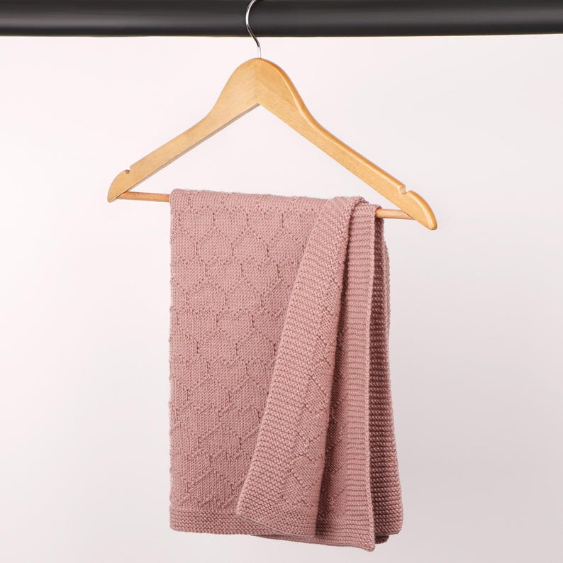 May include: A dusty rose knitted blanket featuring a repeating heart pattern and a wide garter stitch border. The blanket is draped over a light-colored wooden clothes hanger, showcasing the textured knit construction and the soft pink color.