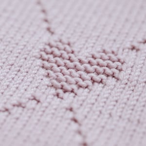 K&ouml;nnte beinhalten: Nahaufnahme eines Strickstoffs in einem weichen, ged&auml;mpften Rosa. Die Textur zeigt ein herzf&ouml;rmiges Design, das mit einer Reihe von Maschen erstellt wurde. Der Hintergrund ist leicht verschwommen, wodurch die Herzform und die Textur des Stoffes hervorgehoben werden.