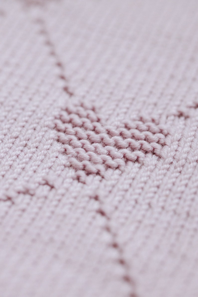 K&ouml;nnte beinhalten: Nahaufnahme eines Strickstoffs in einem weichen, ged&auml;mpften Rosa. Die Textur zeigt ein herzf&ouml;rmiges Design, das mit einer Reihe von Maschen erstellt wurde. Der Hintergrund ist leicht verschwommen, wodurch die Herzform und die Textur des Stoffes hervorgehoben werden.