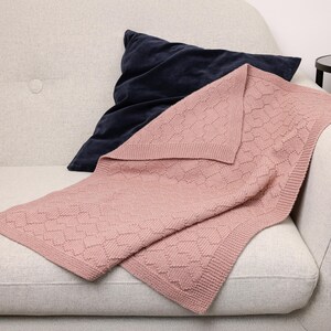 May include: A soft, dusty pink knitted blanket featuring a textured geometric diamond pattern and a wide garter stitch border, layered over a dark navy blue velvet throw pillow. The blanket has a consistent knit construction throughout.
