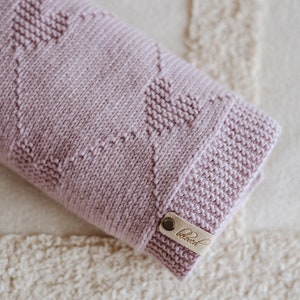 K&ouml;nnte beinhalten: Eine gefaltete, hellviolett gestrickte Decke mit einem Herzmuster. Die Decke hat ein Lederetikett mit dem Wort "beloved". Die Decke liegt gefaltet auf einer cremefarbenen, strukturierten Oberfl&auml;che.