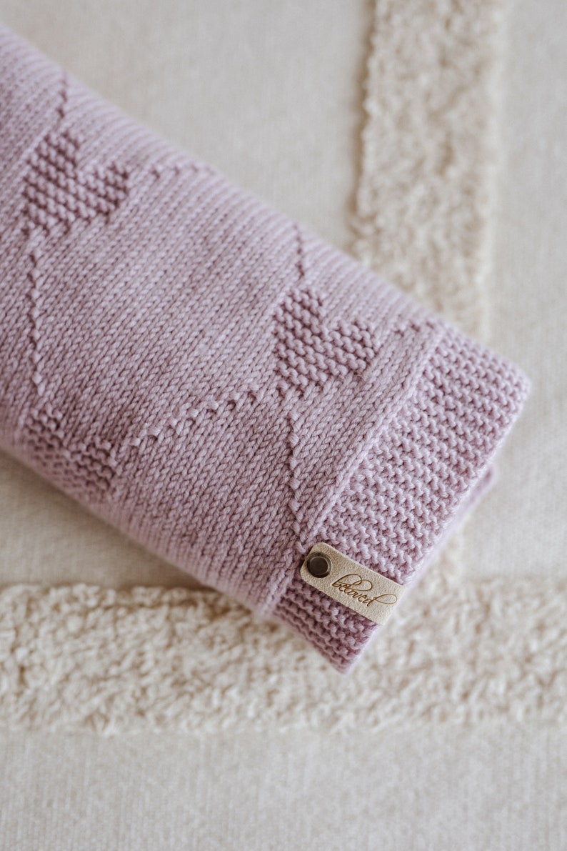 K&ouml;nnte beinhalten: Eine gefaltete, hellviolett gestrickte Decke mit einem Herzmuster. Die Decke hat ein Lederetikett mit dem Wort "beloved". Die Decke liegt gefaltet auf einer cremefarbenen, strukturierten Oberfl&auml;che.