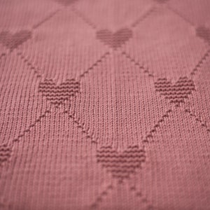 K&ouml;nnte beinhalten: Nahaufnahme einer altrosa gestrickten Decke. Die Decke hat ein sich wiederholendes Muster aus Herzen innerhalb eines Rautenrasters. Die Textur des Strickstoffs ist sichtbar und erzeugt ein weiches und gem&uuml;tliches Aussehen. Das Design ist einfach, aber elegant.