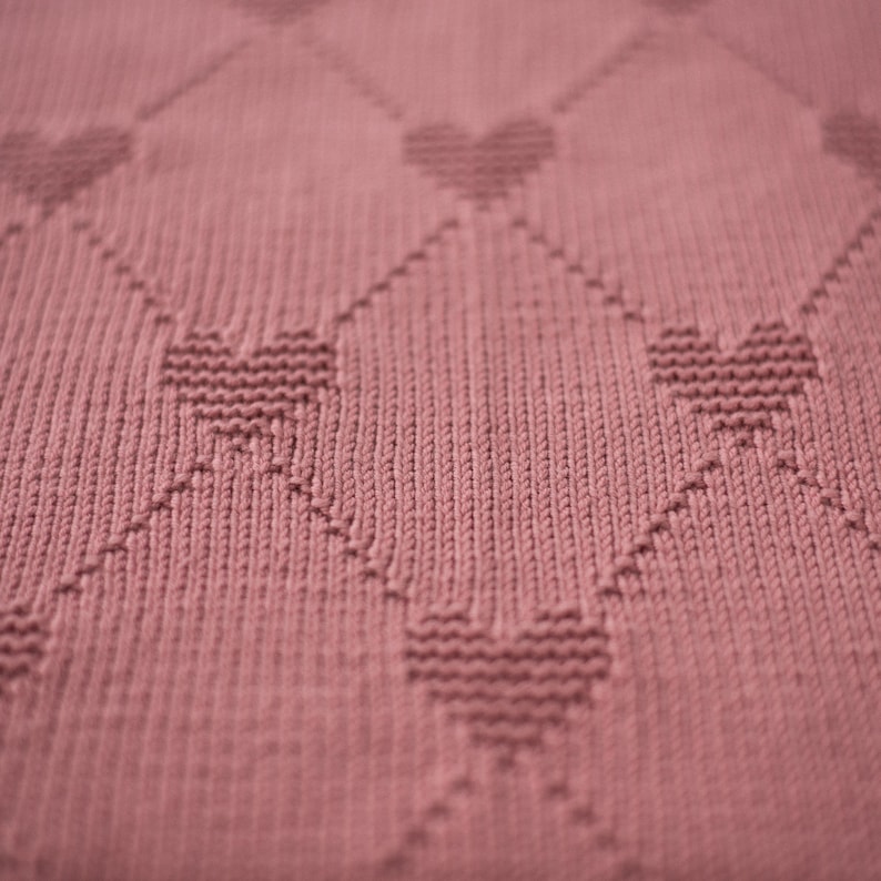 K&ouml;nnte beinhalten: Nahaufnahme einer altrosa gestrickten Decke. Die Decke hat ein sich wiederholendes Muster aus Herzen innerhalb eines Rautenrasters. Die Textur des Strickstoffs ist sichtbar und erzeugt ein weiches und gem&uuml;tliches Aussehen. Das Design ist einfach, aber elegant.