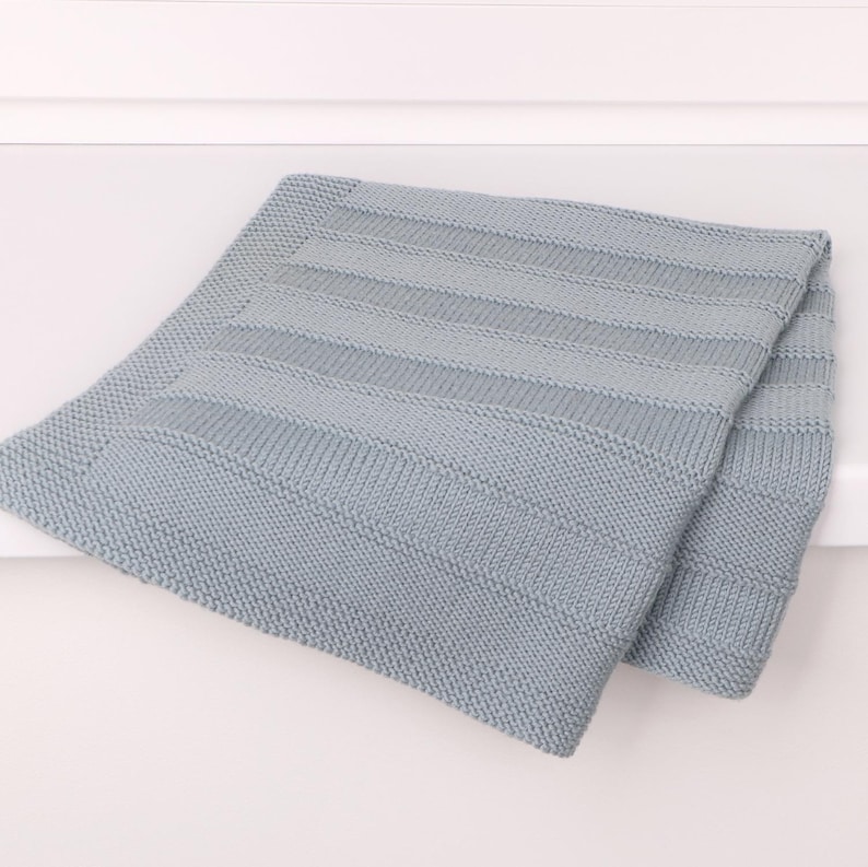 May include: A light blue knitted blanket featuring a textured pattern of alternating horizontal stripes. The design consists of sections of garter stitch borders framing a central area with wider, smooth stockinette stitch bands and narrow ridges.
