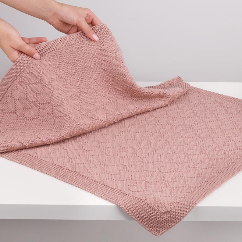 May include: A dusty rose knitted blanket featuring a repeating geometric heart pattern throughout the center and a textured garter stitch border along the edges. The fabric is soft and flexible, showing a consistent knit construction in a muted pink.