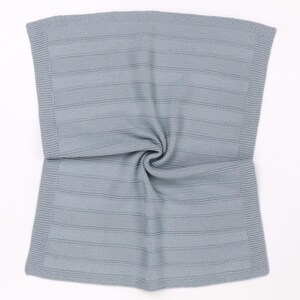 May include: A light gray knitted blanket featuring a textured horizontal striped pattern throughout the center. The edges of the rectangular blanket are finished with a distinct garter stitch border, creating a uniform and structured appearance.