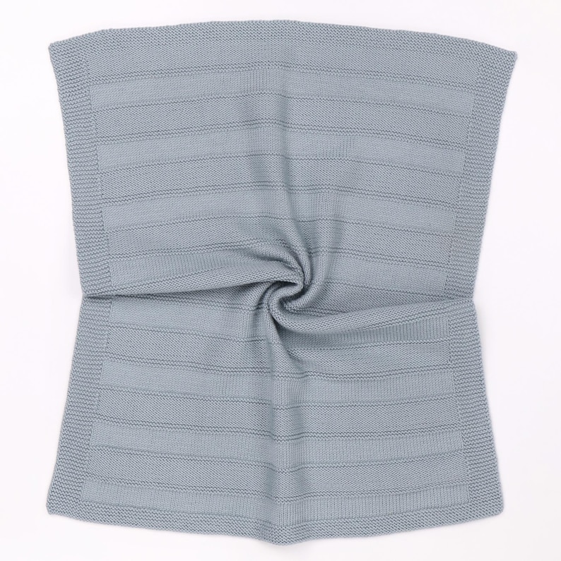 May include: A light gray knitted blanket featuring a textured horizontal striped pattern throughout the center. The edges of the rectangular blanket are finished with a distinct garter stitch border, creating a uniform and structured appearance.