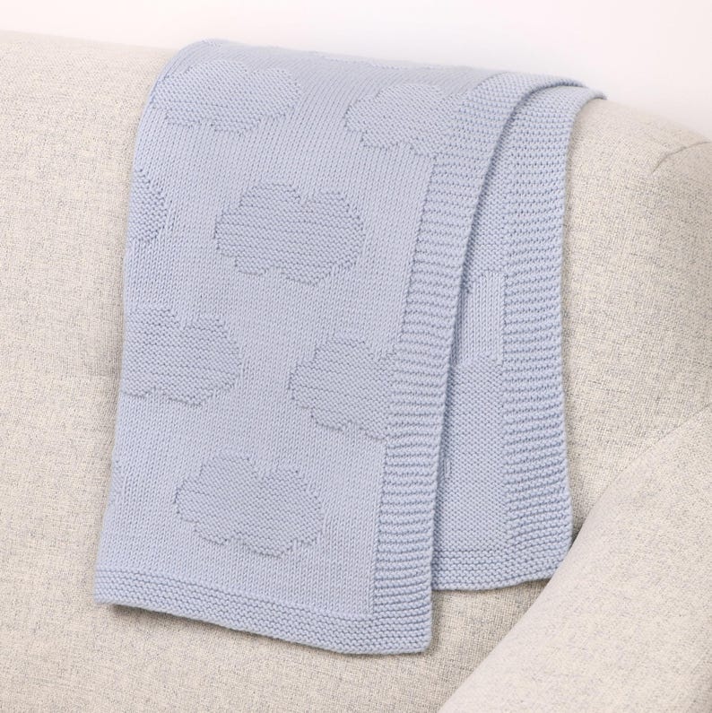 May include: A light blue knitted blanket featuring a repeating pattern of textured clouds. The blanket has a wide, ribbed border along the edges and a consistent knit construction throughout, showcasing a soft, uniform color and tactile design.