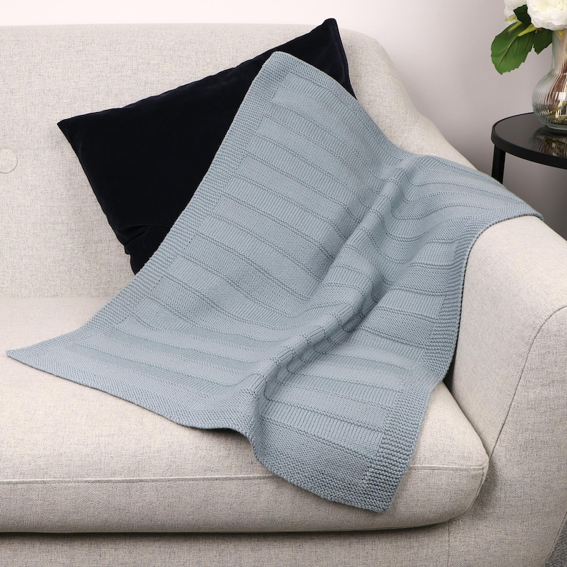 May include: A light blue knitted blanket featuring a textured pattern of alternating horizontal stripes and a solid border. The fabric has a consistent weave throughout, showcasing a soft appearance with a defined edge finish and a rectangular shape.