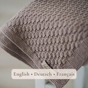 Könnte beinhalten: Eine taupefarbene Strickdecke mit einem texturierten, sich wiederholenden Reliefmuster. Der Stoff weist ein gleichmäßiges, dichtes Maschenbild auf. An der Ecke ist ein kleines, helles, rechteckiges Stoffetikett mit kursivem Text angebracht.