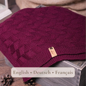 May include: A deep burgundy knitted blanket featuring a textured geometric diamond pattern throughout. A small, rectangular tan leather tag with a metallic rivet is attached to the corner, displaying the text English • Deutsch • Français.