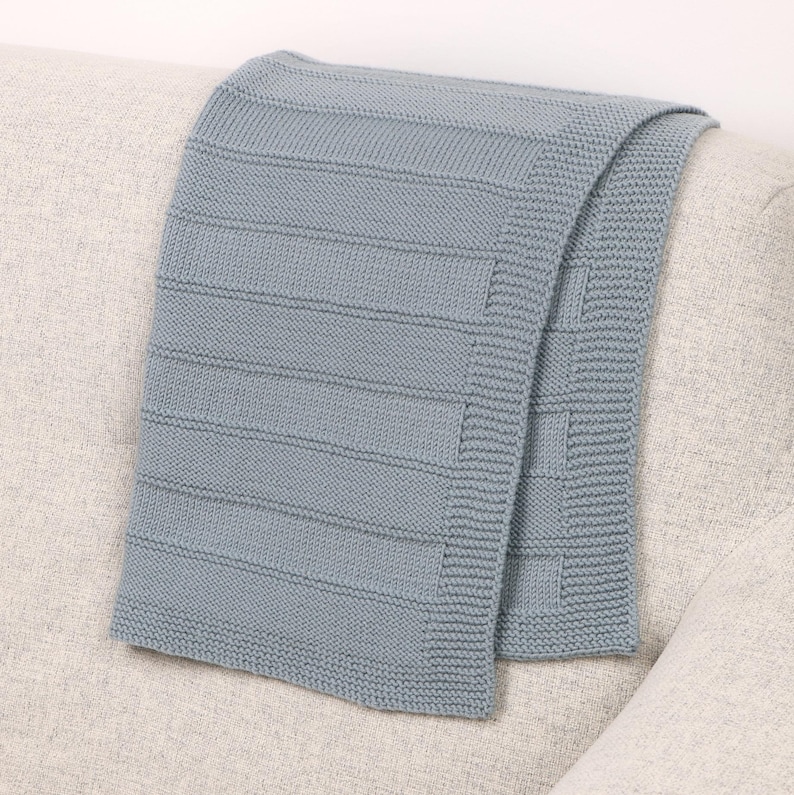 May include: A light blue knitted throw blanket featuring a textured pattern of horizontal rectangular blocks separated by narrow recessed lines, with a wide garter stitch border along the edges, creating a uniform and structured appearance.