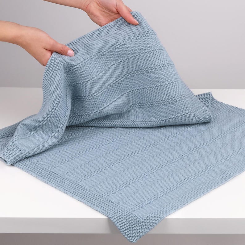 May include: A light blue knitted blanket featuring a textured garter stitch border and a central panel with alternating vertical stripes of garter stitch and smooth stockinette stitch. The fabric is soft and flexible with a uniform light blue color.